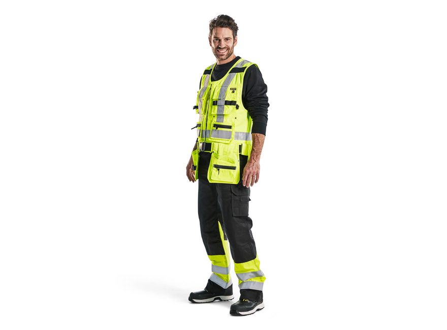 Man wearing yellow hi-vis tool vest over black long-sleeve shirt and dark work trousers, on a white background